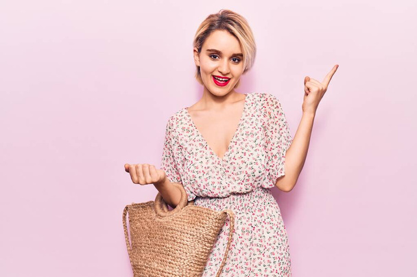 Young beautiful blonde woman wearing casual clothes holding wicker bag smiling happy pointing with hand and finger to the side  - Photo, Image