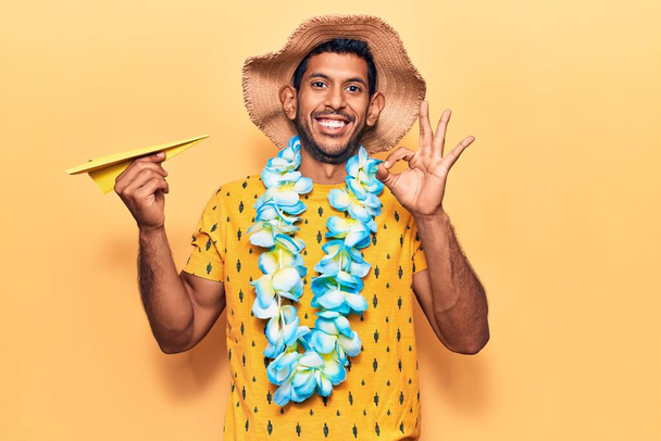 Νεαρός Λατίνος άνδρας φορώντας καλοκαιρινό καπέλο και hawaiian lei κρατώντας χάρτινο αεροπλάνο κάνει ok υπογράψει με τα δάχτυλα, χαμογελώντας φιλικό gesturing εξαιρετικό σύμβολο  - Φωτογραφία, εικόνα