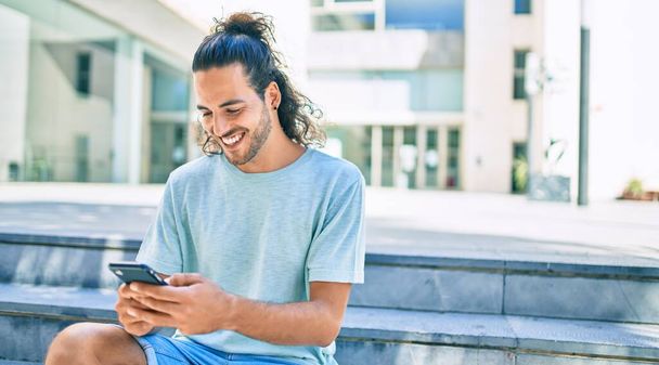 Mladý hispánec muž usměvavý šťastný pomocí smartphone ve městě. - Fotografie, Obrázek
