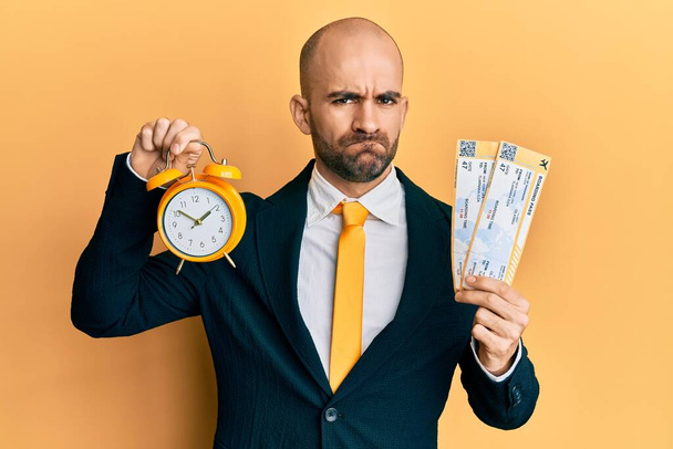 Young hispanic business man holding boarding pass and alarm clock skeptic and nervous, frowning upset because of problem. negative person.  - 写真・画像