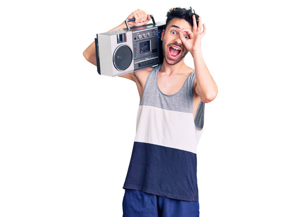 Młody Hiszpan trzyma boombox, słuchając muzyki uśmiechnięty szczęśliwy robi znak ok z ręką na oku patrząc przez palce  - Zdjęcie, obraz