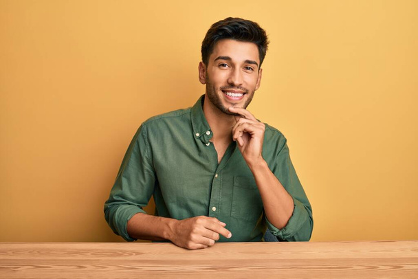 Jonge knappe man in casual kleding zittend op de tafel glimlachend zelfverzekerd kijkend naar de camera met gekruiste armen en hand op kin. positief denken.  - Foto, afbeelding
