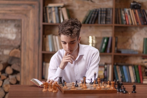 Подросток играет в шахматы сам с собой, мальчик сидит за большим деревянным столом и обдумывает шахматные ходы в игре, белые и черные шахматные фигуры, шахматы и мат. - Фото, изображение