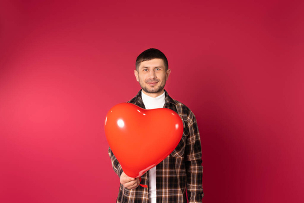 Portrait of a young brunette guy looking to camera, holding a heart-shaped balloon on a red background. Holiday concept. High quality photo - Fotó, kép