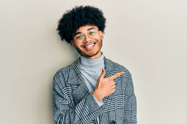 Молодий афроамериканець з волоссям афроамериканського кольору в келихах повсякденного одягу, що посміхаються з веселим наконечником з рукою і пальцем на бік.  - Фото, зображення