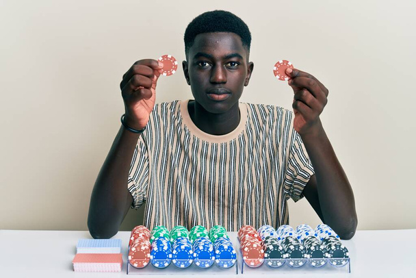 Nuori afrikkalainen amerikkalainen mies pelaa pokeria tilalla pelimerkkejä rento vakava ilme kasvoilla. yksinkertainen ja luonnollinen kameraan katsominen.  - Valokuva, kuva