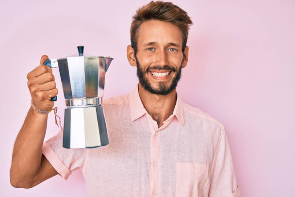 Knappe blanke man met baard houdt Italiaanse koffiezetapparaat kijken positief en gelukkig staan en glimlachen met een zelfverzekerde glimlach tonen tanden  - Foto, afbeelding