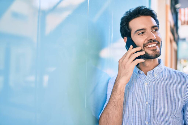 Молодий іспаномовний чоловік посміхається щасливим розмовляти на смартфоні, спираючись на стіну
 - Фото, зображення
