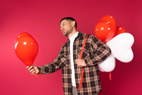 A young man holds out a red heart-shaped balloon and holds a bunch of white and red balloons behind his back. High quality photo - Fotó, kép