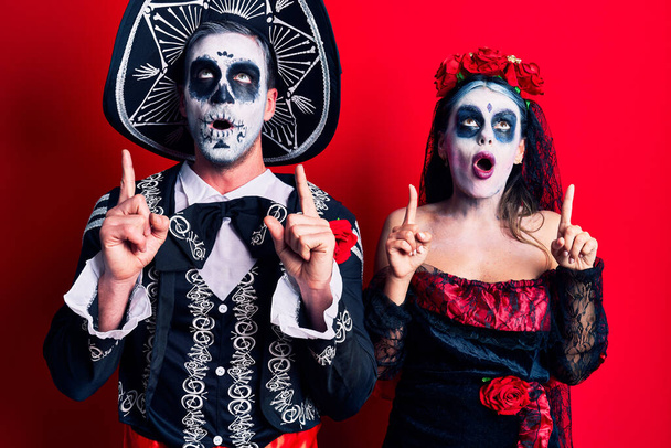 Mladý pár v mexickém dni mrtvého kostýmu nad červeným užaslý a překvapený při pohledu nahoru a ukazuje prsty a zvednutými pažemi.  - Fotografie, Obrázek