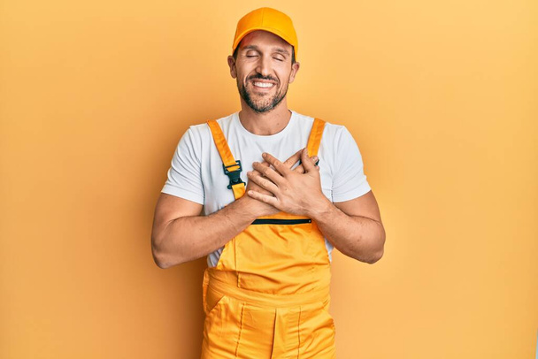 Young handsome man wearing handyman uniform over yellow background smiling with hands on chest with closed eyes and grateful gesture on face. health concept.  - Photo, Image