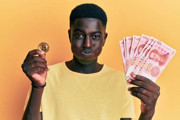 Young african american man holding yuan chinese banknotes and bitcoin puffing cheeks with funny face. mouth inflated with air, catching air.  - Foto, immagini