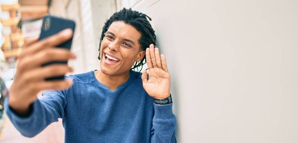 若いアフリカ系アメリカ人の男が壁にもたれてスマートフォンを使ってビデオ通話をする. - 写真・画像