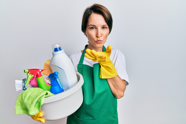 Young brunette woman with short hair wearing apron holding cleaning products looking at the camera blowing a kiss with hand on air being lovely and sexy. love expression.  - Photo, Image