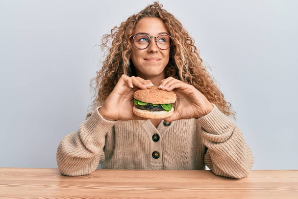 Schöne kaukasische Teenager-Mädchen essen einen leckeren klassischen Burger lächelnd zur Seite schauen und wegstarren denken.  - Foto, Bild