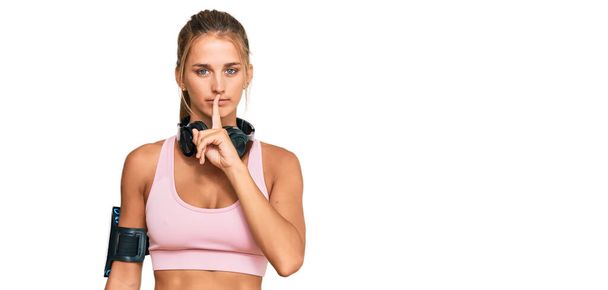 Young blonde woman wearing gym clothes and using headphones asking to be quiet with finger on lips. silence and secret concept.  - Foto, Imagen