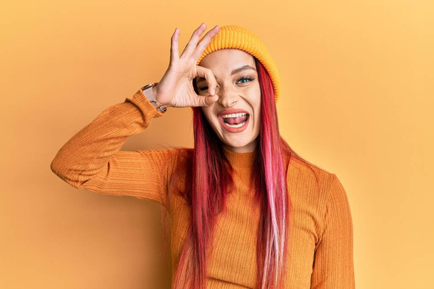 Jovem mulher caucasiana vestindo boné de lã sorrindo feliz fazendo ok sinal com a mão no olho olhando através dos dedos  - Foto, Imagem