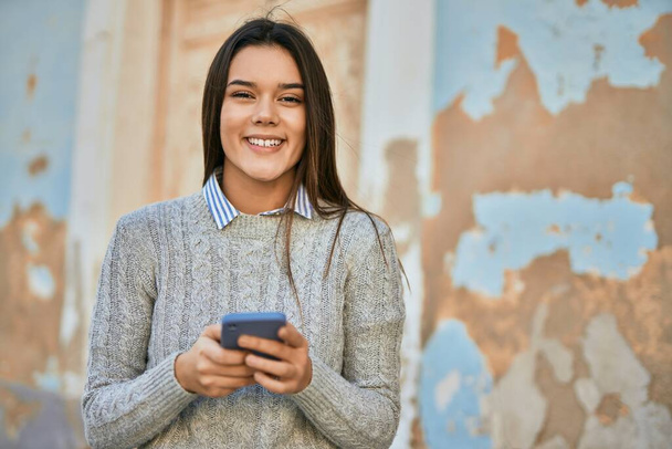 Nuori latino tyttö hymyilee onnellinen käyttäen älypuhelin kaupungin. - Valokuva, kuva
