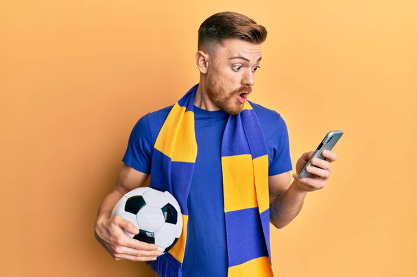 Young redhead man wearing hooligan scarf and using smartphone afraid and shocked with surprise and amazed expression, fear and excited face.  - Photo, Image