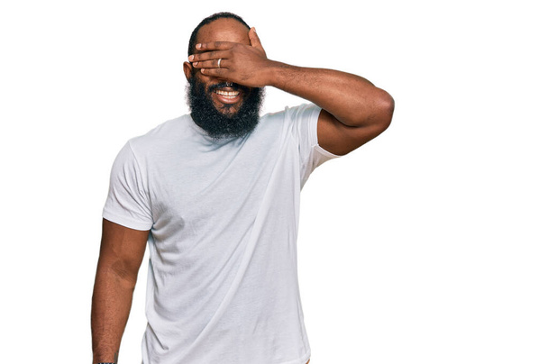 Young african american man wearing casual white tshirt smiling and laughing with hand on face covering eyes for surprise. blind concept.  - Photo, Image