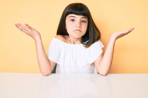 Young little girl with bang wearing casual clothes sitting on the table clueless and confused with open arms, no idea and doubtful face.  - Photo, Image