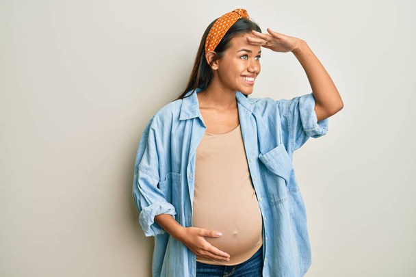 Красива іспаномовна жінка очікує на дитину, торкаючись вагітного живота дуже щаслива і посміхається, дивлячись далеко за голову. концепція пошуку
.  - Фото, зображення