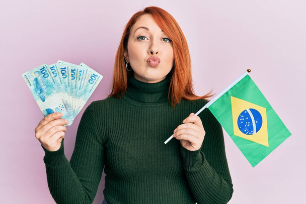Krásná zrzka žena drží 100 brazilských pravých bankovek a brazilské vlajky při pohledu na fotoaparát foukání polibek je krásná a sexy. výraz lásky.  - Fotografie, Obrázek