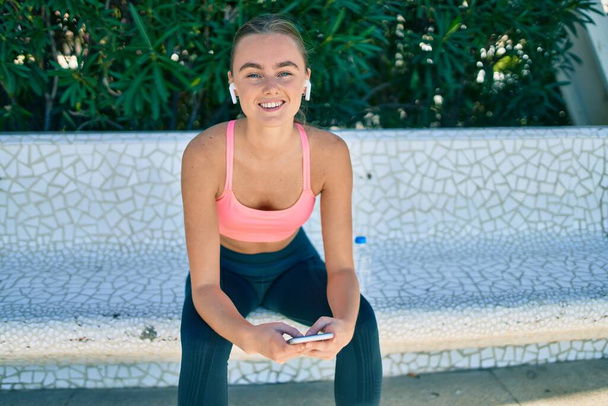 Young blonde sportswoman doing exercise using smartphone sitting on the bench at the park. - Foto, Imagem