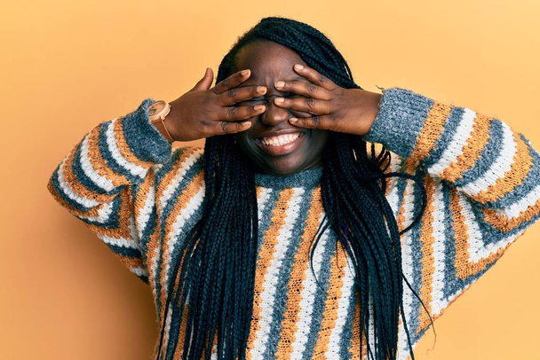Young black woman with braids wearing casual winter sweater covering eyes with hands smiling cheerful and funny. blind concept.  - Fotografie, Obrázek