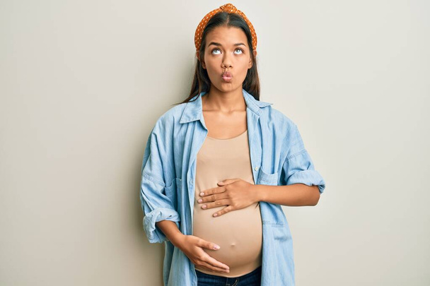 Gyönyörű spanyol nő babát vár, terhes hasat érintve halfejet vágva ajkakkal, őrült és komikus gesztussal. Vicces kifejezés..  - Fotó, kép