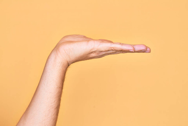 Hand of caucasian young man showing fingers over isolated yellow background with flat palm presenting product, offer and giving gesture, blank copy space - Foto, afbeelding