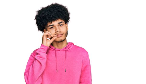 Jeune homme afro-américain aux cheveux afro portant un sweat-shirt rose décontracté bouche et lèvres fermées comme zip avec les doigts. secret et silencieux, tabou parlant  - Photo, image