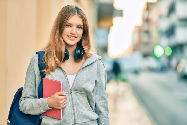 Beautiful caucasian student teenager using headphones holding book at the city. - Fotó, kép