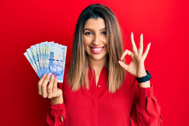 Hermosa mujer morena sosteniendo billetes de 100 rand sudafricanos haciendo signo ok con los dedos, sonriendo gesto amistoso excelente símbolo  - Foto, Imagen