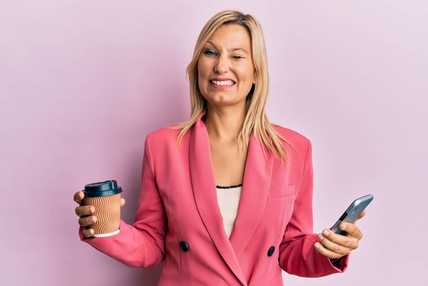 Beautiful middle age blonde woman using smartphone and drinking a cup of coffee winking looking at the camera with sexy expression, cheerful and happy face.  - Fotoğraf, Görsel