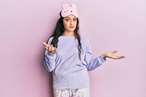 Beautiful middle eastern woman wearing sleep mask and pajama clueless and confused expression with arms and hands raised. doubt concept.  - Photo, Image
