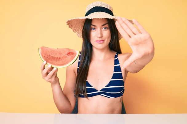 Young beautiful caucasian woman wearing bikini and hat holding slice of watermelon with open hand doing stop sign with serious and confident expression, defense gesture  - Φωτογραφία, εικόνα