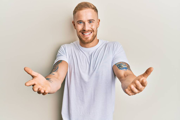 Young caucasian man wearing casual white tshirt looking at the camera smiling with open arms for hug. cheerful expression embracing happiness.  - Photo, Image