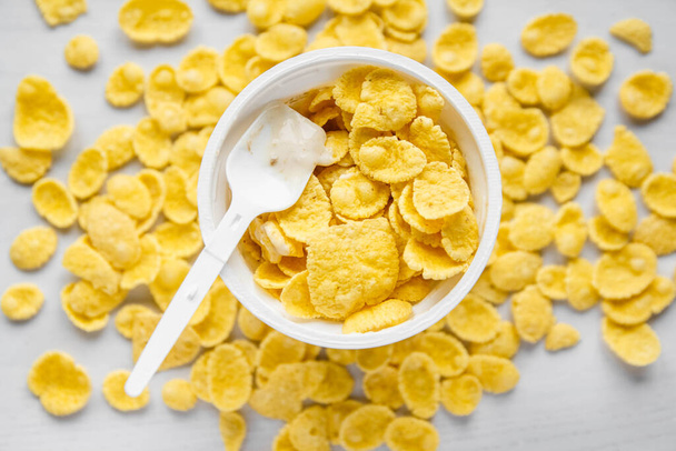 Cornflakes mit Joghurt in weißer Schüssel mit Löffel auf weißem Holzhintergrund. Ansicht von oben. Kopieren, leerer Raum für Text. - Foto, Bild