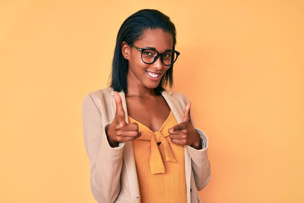 Mladá afro-americká žena, oblečená do obchodního oblečení, ukazuje prstem na kameru se šťastnou a zábavnou tváří. dobrá energie a vibrace.  - Fotografie, Obrázek