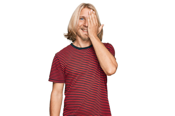 Blanke man met blond lang haar met casual gestreept t-shirt over één oog met de hand, zelfverzekerde glimlach op het gezicht en verrassende emotie.  - Foto, afbeelding