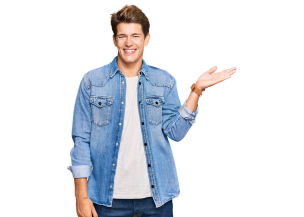 Handsome caucasian man wearing casual denim jacket smiling cheerful presenting and pointing with palm of hand looking at the camera.  - Photo, Image