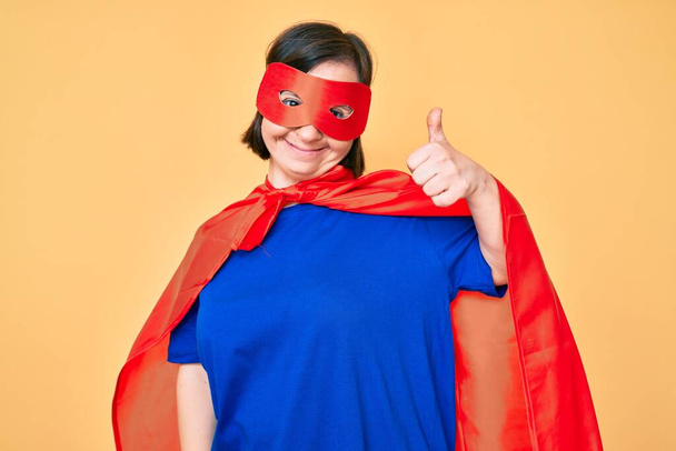 Down sendromlu esmer kadın süper kahraman kostümü giyiyor mutlu ve pozitif gülümsüyor, başparmağıyla mükemmel ve onay işareti yapıyor.  - Fotoğraf, Görsel