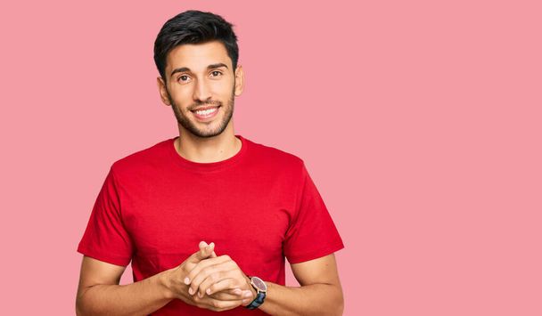 Ein junger gutaussehender Mann in lässigem rotem T-Shirt mit zusammengelegten Händen und Daumen, der entspannt und fröhlich lächelt. Erfolg und Optimismus  - Foto, Bild