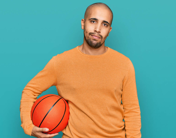 Hispanic adult man holding basketball ball thinking attitude and sober expression looking self confident  - 写真・画像