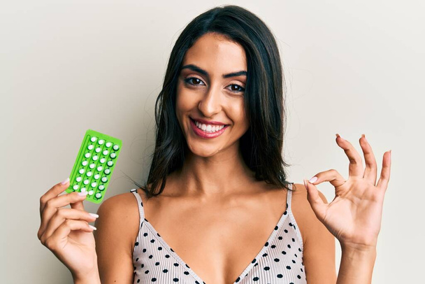 Belle femme hispanique tenant des pilules contraceptives faisant signe ok avec les doigts, sourire geste amical excellent symbole  - Photo, image