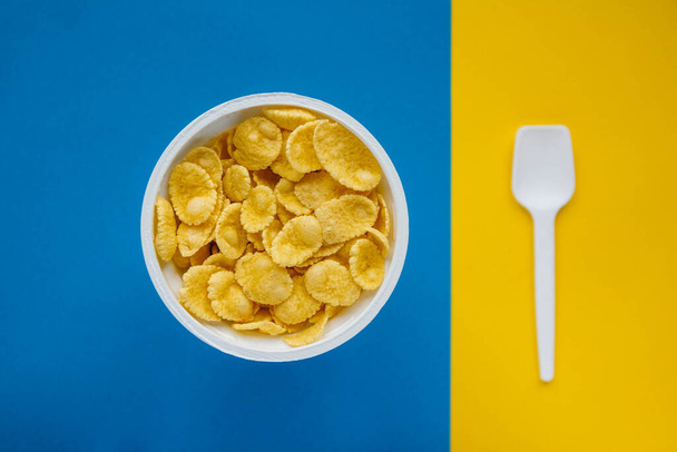Copos de maíz en tazón blanco con cuchara sobre fondo azul y amarillo. Vista superior. Copiar, espacio vacío para texto. - Foto, Imagen