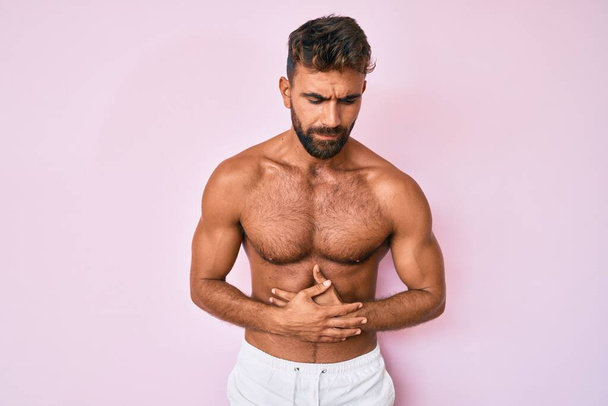 Jonge Spaanse man die zonder shirt staat met de hand op de maag omdat indigestie, pijnlijke ziekte zich onwel voelt. pijnconcept.  - Foto, afbeelding