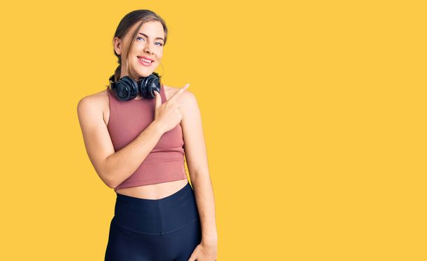 Beautiful caucasian young woman wearing gym clothes and using headphones cheerful with a smile on face pointing with hand and finger up to the side with happy and natural expression  - Fotografie, Obrázek