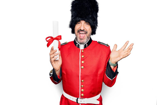 Мужчина среднего возраста красивый Уэльс охранник носить традиционную униформу проведение диплом степени празднования достижений с счастливой улыбкой и выражение победителя с поднятой рукой - Фото, изображение